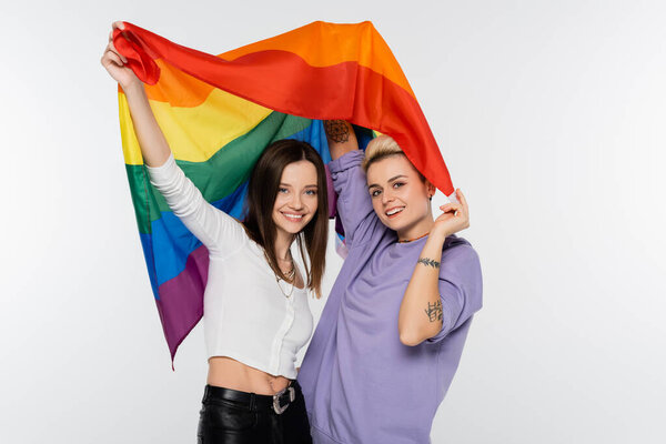 happy and trendy lesbians looking at camera while holding lgbtq flag isolated on grey