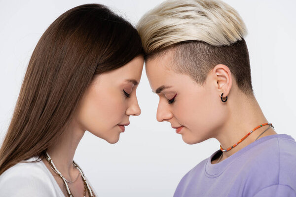 side view of brunette and blonde lesbian couple standing face to face with closed eyes isolated on grey