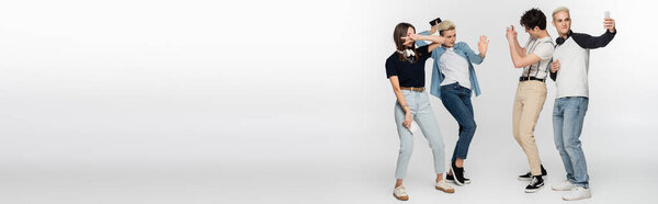 young woman showing victory sign and dancing near friends taking photo on smartphones on grey background, banner