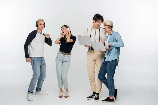 stock image smiling couple using laptops near cheerful friends dancing in wireless headphones on grey background