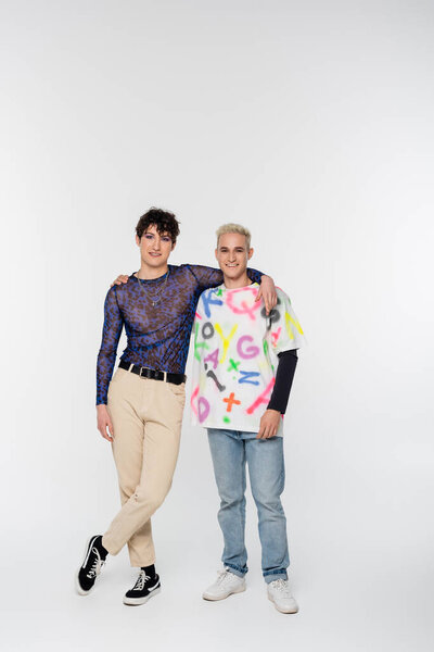 full length of young and joyful queer person with gay man hugging and looking at camera on grey background
