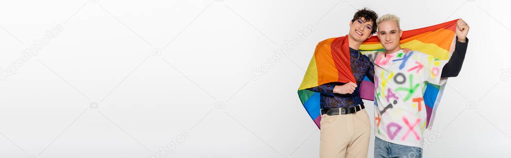trendy queer person and blonde gay man with lgbtq flag smiling at camera isolated on grey, banner