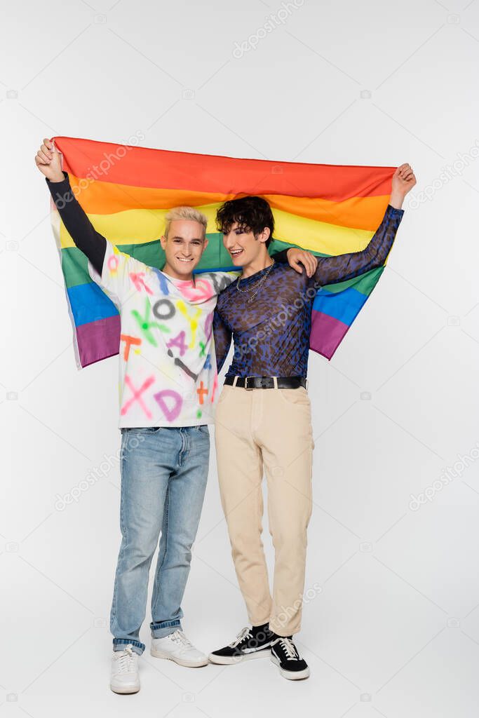 full length of gay man and nonbinary person holding lgbtq flag on grey background