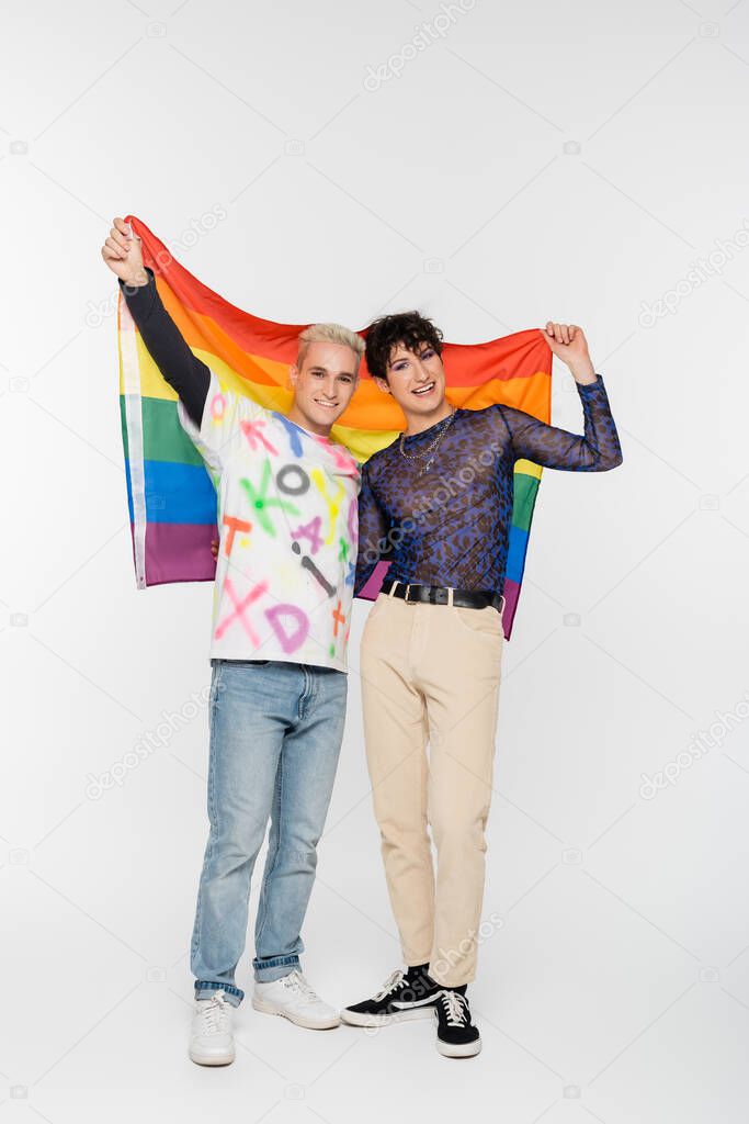 full length of gay man and nonbinary person standing with lgbtq flag on grey background