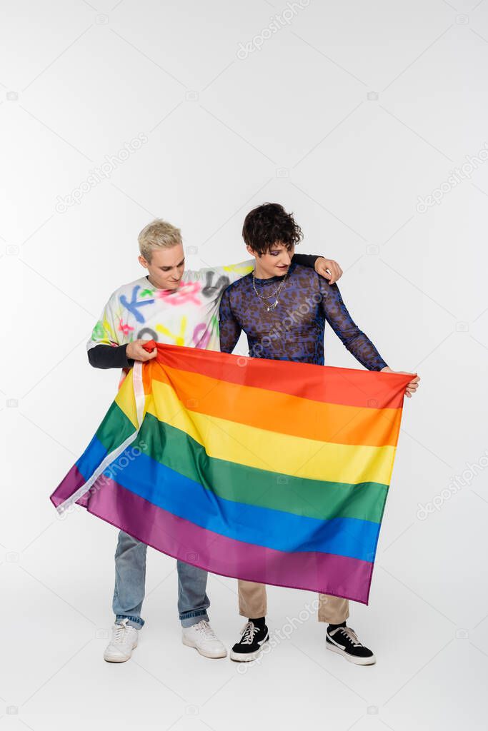 blonde gay man and brunette nonbinary person standing with lgbtq flag on grey background