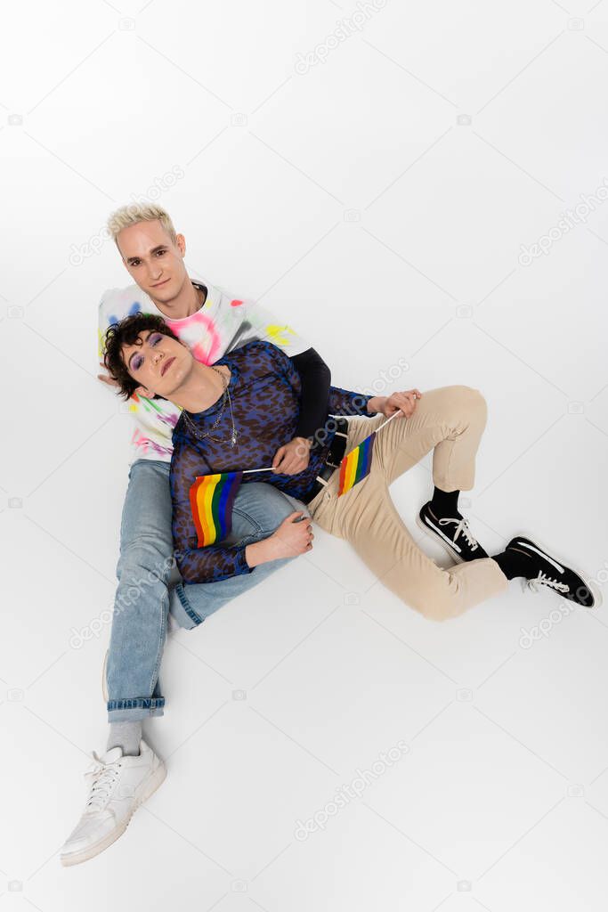 top view of stylish gay man and queer person sitting with lgbtq flags on grey background