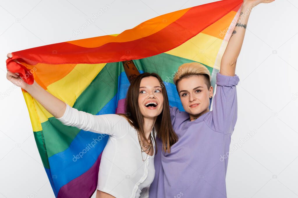 cheerful young lesbians holding lgbtq flag isolated on grey