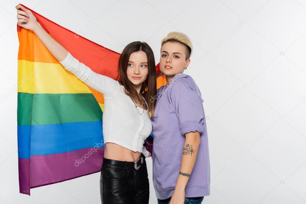 tattooed lesbian woman looking at camera near girlfriend with lgbtq flag isolated on grey