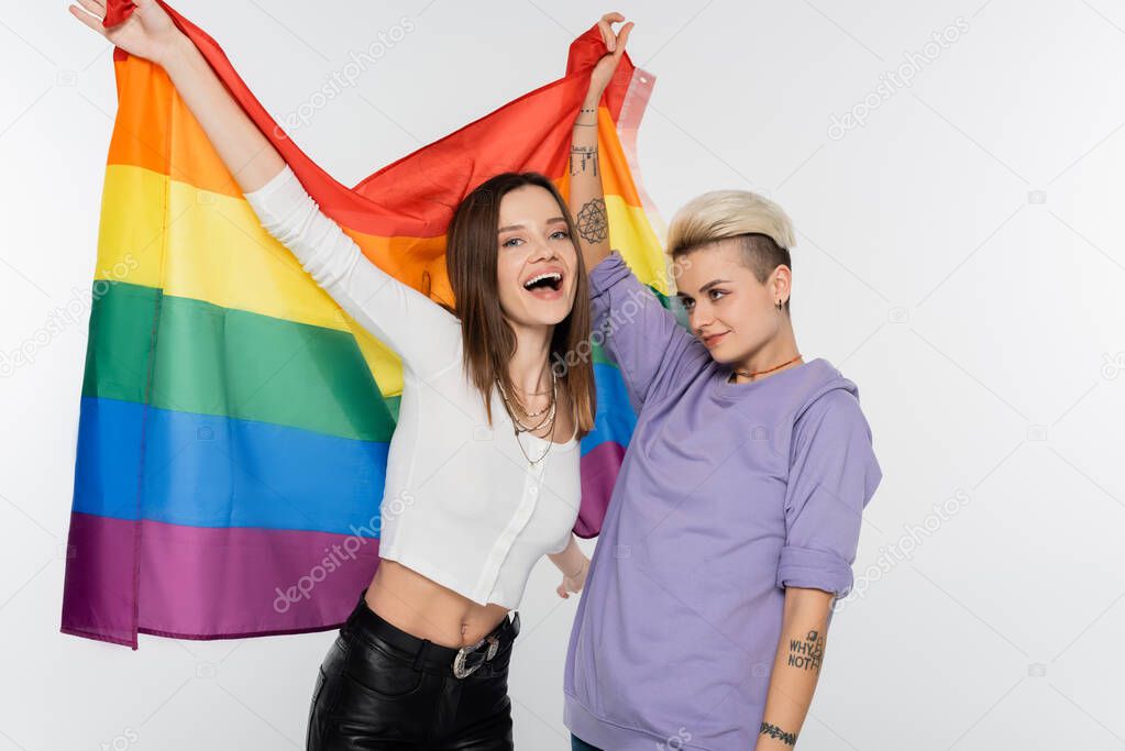tattooed lesbian woman with excited girlfriend holding lgbtq flag isolated on grey