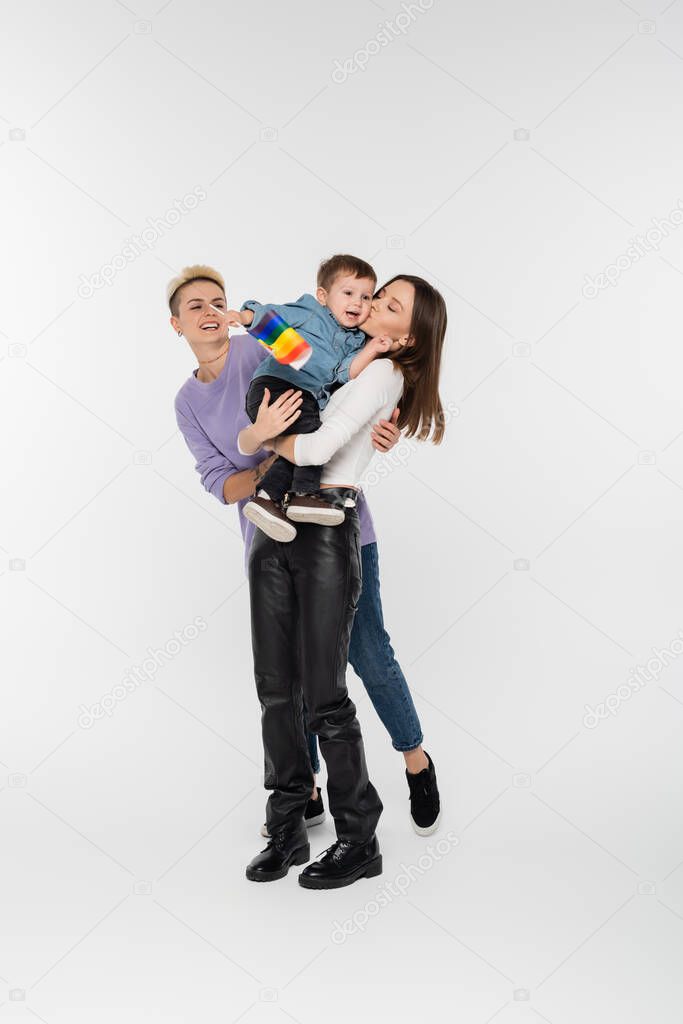 full length of happy lesbians holding boy with lgbtq flag on grey background