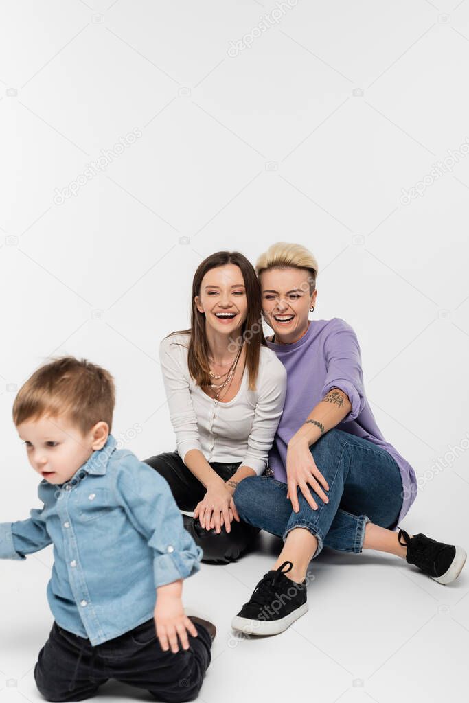 cheerful lesbian couple laughing near toddler son on grey background