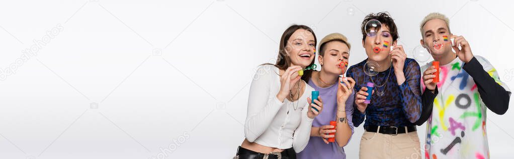 stylish people of different identity and with lgbtq flags on faces blowing soap bubbles isolated on grey, banner