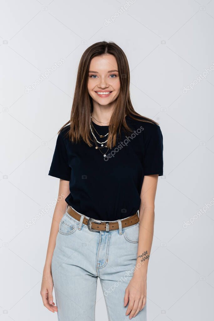 young brunette woman in black t-shirt and necklaces smiling at camera isolated on grey