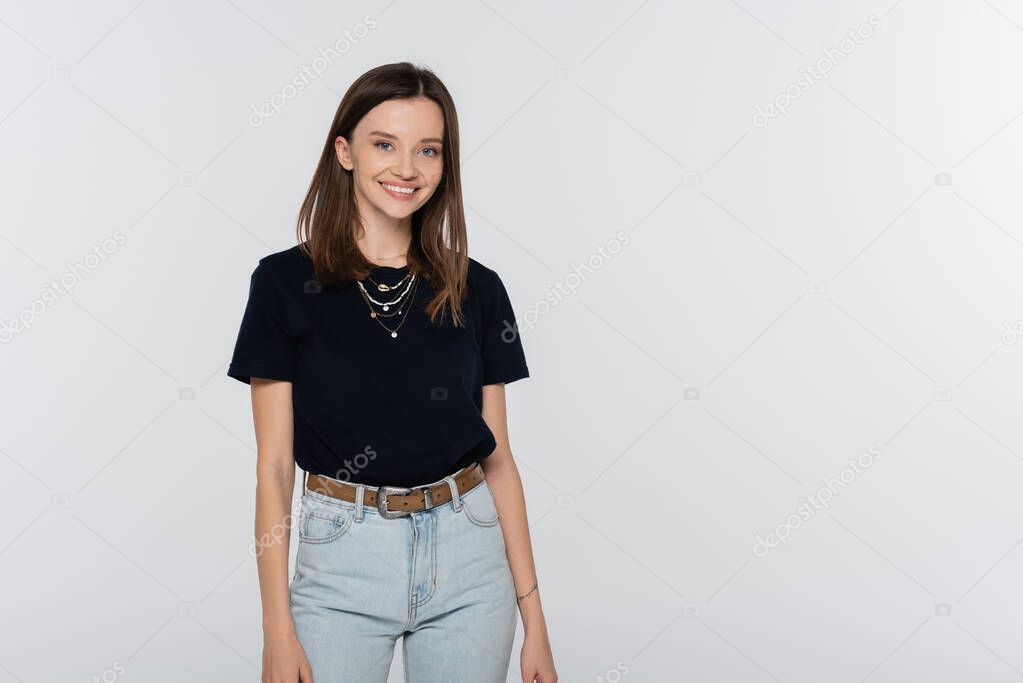 pretty and happy woman in black t-shirt and jeans smiling at camera isolated on grey