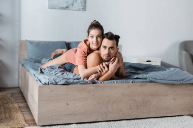 Genç ve seksi çift kameraya gülümserken gri yatak örtüsüne sarılıyorlar.