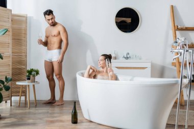 Gülümseyen bir kadın, küvette, erkek arkadaşının yanında iç çamaşırlı, elinde şampanya bardağıyla dikiliyor.