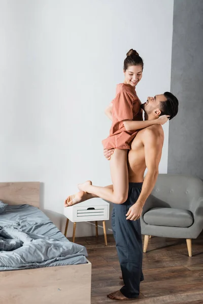 muscular man in pajama pants holding smiling girlfriend in bedroom