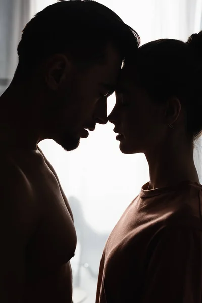side view of silhouettes of woman in t-shirt and shirtless man standing face to face with closed eyes