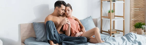 shirtless man in pajama pants holding hands with girlfriend in t-shirt while sitting on bed, banner