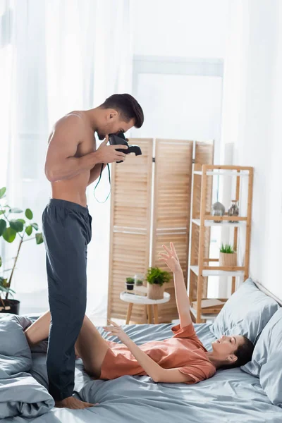 side view of shirtless man in pajama pants taking photo of woman lying on grey bedding