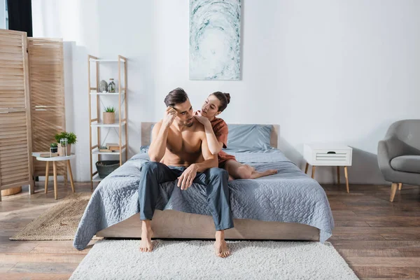 Young Woman Embracing Shirtless Boyfriend Sitting Pajama Pants Spacious Bedroom — Stock Photo, Image