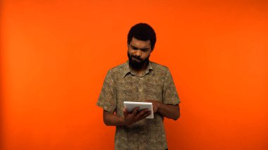 puzzled african american young man with beard holding digital tablet on orange background  clipart
