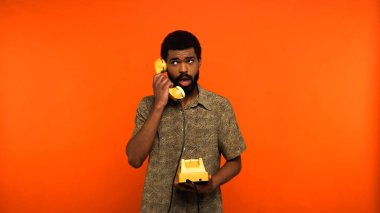 Sakallı genç Afrikalı Amerikalı adam turuncu arka planda sarı retro telefondan konuşuyor. 