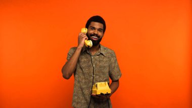 Sakallı neşeli Afrikalı Amerikalı adam turuncu arka planda retro telefondan konuşuyor. 