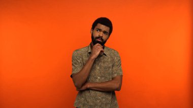 thoughtful african american man in shirt looking away while standing on orange background  clipart