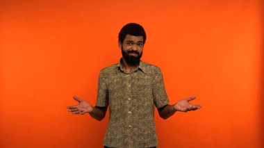 uncertain african american man in shirt looking at camera while gesturing on orange background  clipart