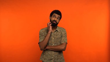 Düşünceli Afrikalı Amerikalı genç adam gözlerini kaçırıyor ve turuncu arka planda sakal çiziyor.