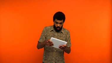 Şok olmuş Afrikalı Amerikalı sakallı genç adam turuncu arka planda dijital tablet tutuyor. 