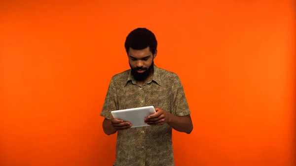 有胡子的非洲裔美国年轻人在橙色背景下玩游戏时使用数码平板电脑 — 图库照片