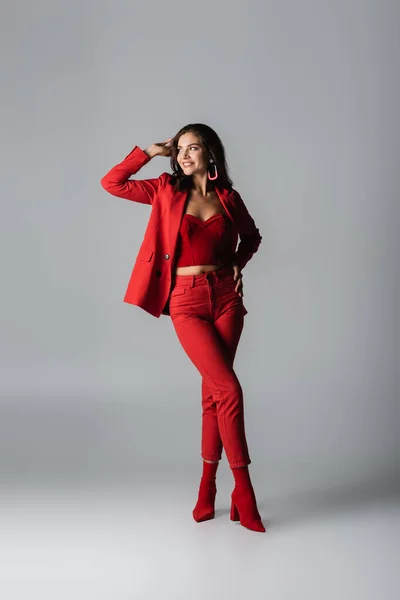 Longitud completa de la joven feliz mujer en botas rojas y traje posando en gris - foto de stock