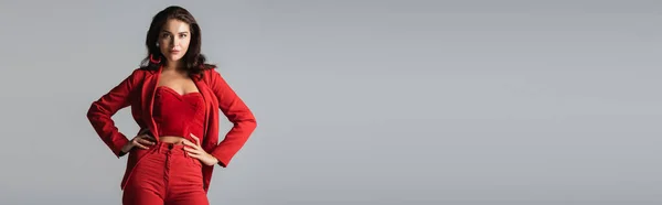 Jeune femme tendance en costume rouge et boucles d'oreilles posant les mains sur les hanches isolées sur gris, bannière — Photo de stock