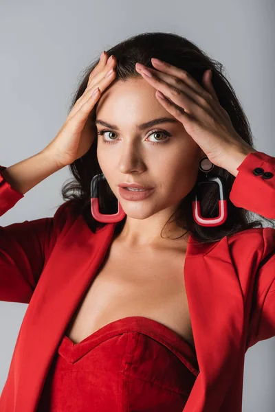 Jeune femme à la mode en costume rouge et boucles d'oreilles regardant la caméra isolée sur gris — Photo de stock