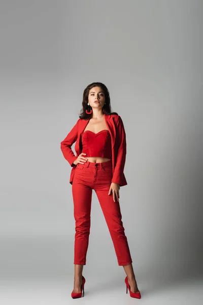 Полная длина молодой стильной женщины на красных каблуках и в костюме, позирующем с рукой на бедре на сером — стоковое фото