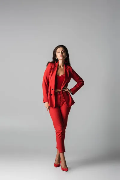 Pleine longueur de jeune femme confiante en talons rouges et costume posant avec la main sur la hanche sur gris — Photo de stock