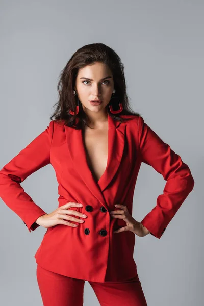 Stylish woman in red suit posing and looking at camera while posing with hands on hips isolated on grey — Stock Photo