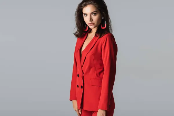 Femme à la mode en costume rouge posant et regardant la caméra isolée sur gris — Photo de stock