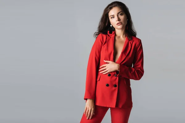 Mujer joven con estilo en traje rojo posando y mirando a la cámara aislada en gris - foto de stock