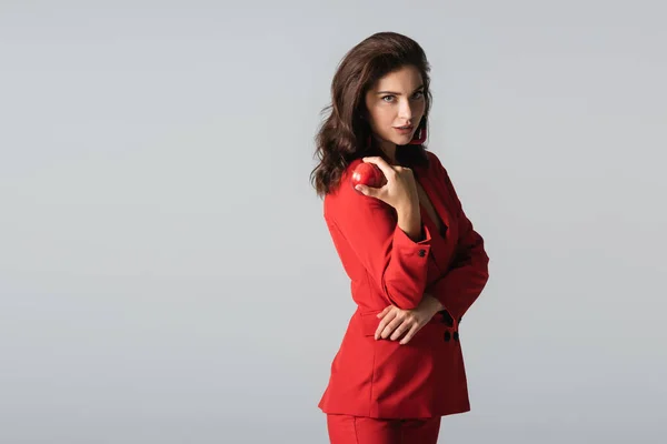 Mujer con estilo en traje rojo posando con manzana aislada en gris - foto de stock