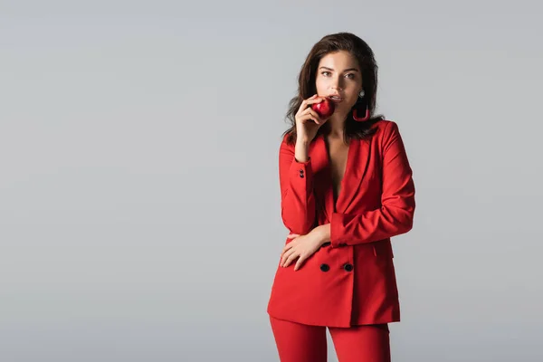 Модная молодая женщина в красном костюме позирует со свежим яблоком, изолированным на сером — стоковое фото