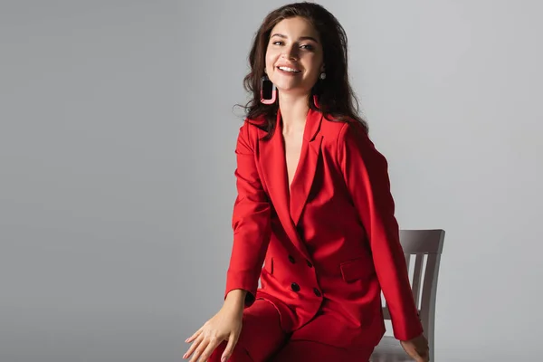 Feliz joven en traje rojo posando mientras está sentado en silla blanca aislado en gris - foto de stock