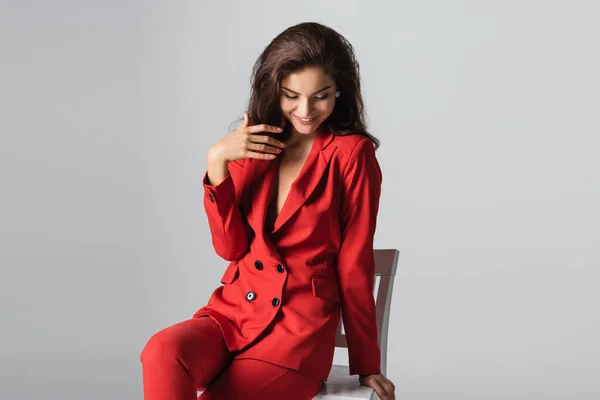 Fröhliche junge Frau im roten Anzug posiert auf weißem Stuhl isoliert auf grau — Stockfoto