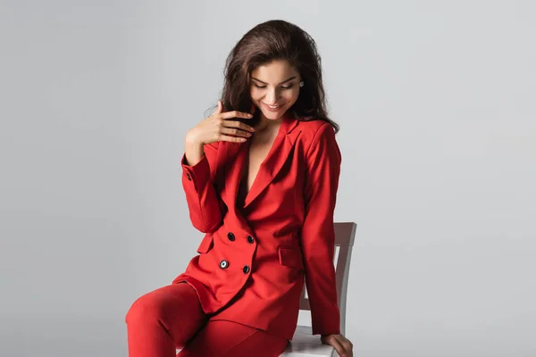 Alegre joven en traje rojo posando sentado en silla blanca aislado en gris - foto de stock