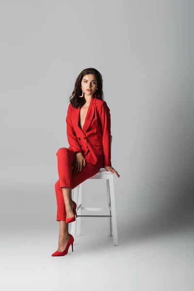 Повна довжина модної жінки в червоному стильному костюмі позує сидячи на білому стільці на сірому — стокове фото