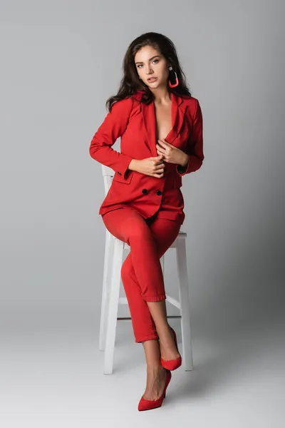 Longitud completa de la mujer joven sexy en traje rojo posando mientras está sentado en la silla blanca en gris - foto de stock