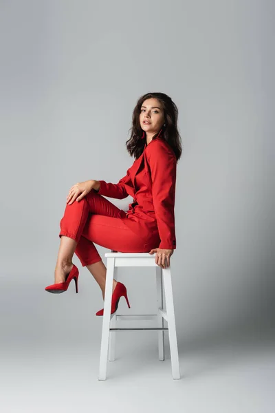 Pleine longueur de jeune femme à la mode en costume rouge assis sur chaise blanche sur gris — Photo de stock
