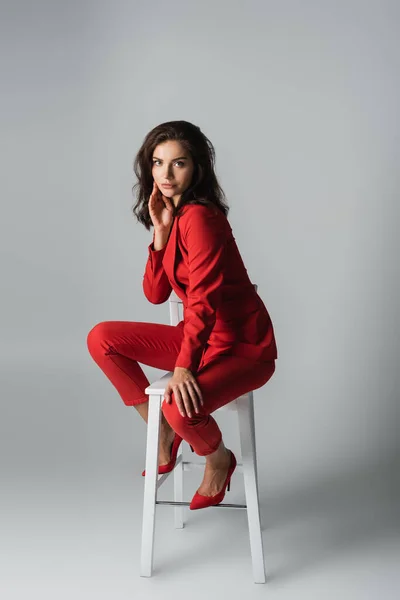 Longitud completa de la mujer de moda en traje rojo sentado en silla blanca en gris - foto de stock